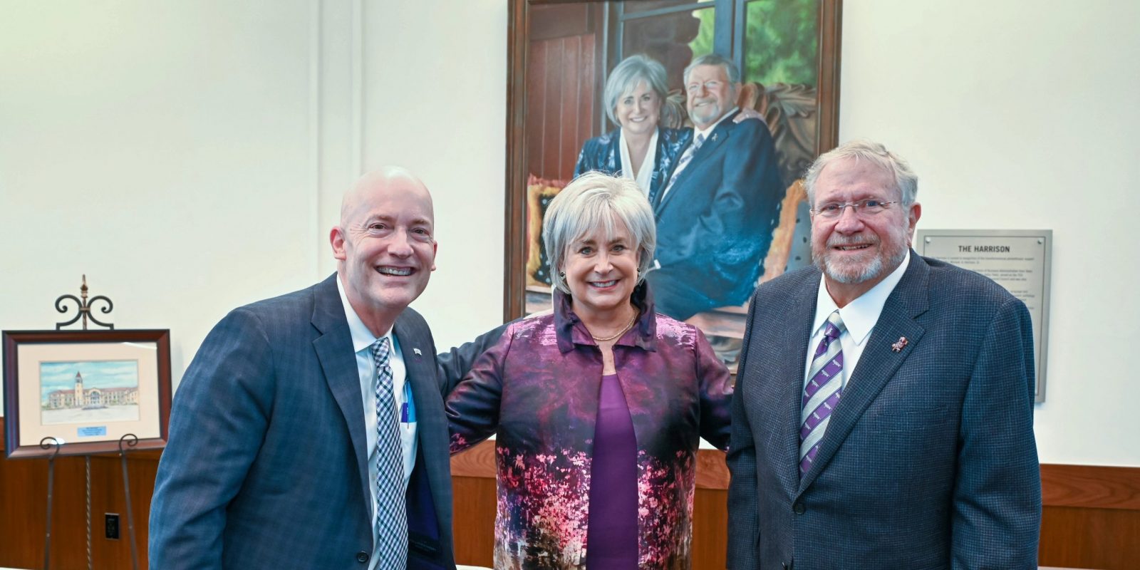 Mike Harrison and his wife, Brenda, pose with TCU Chancellor Emeritus Victor J. Boschini Jr. beneath a commissioned portrait of the couple inside The Harrison building on Texas Christian University’s campus. A watercolor illustration or painting of The Harrison building rests on an easel to the left.