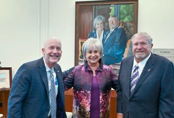 Mike Harrison and his wife, Brenda, pose with TCU Chancellor Emeritus Victor J. Boschini Jr. beneath a commissioned portrait of the couple inside The Harrison building on Texas Christian University’s campus. A watercolor illustration or painting of The Harrison building rests on an easel to the left.