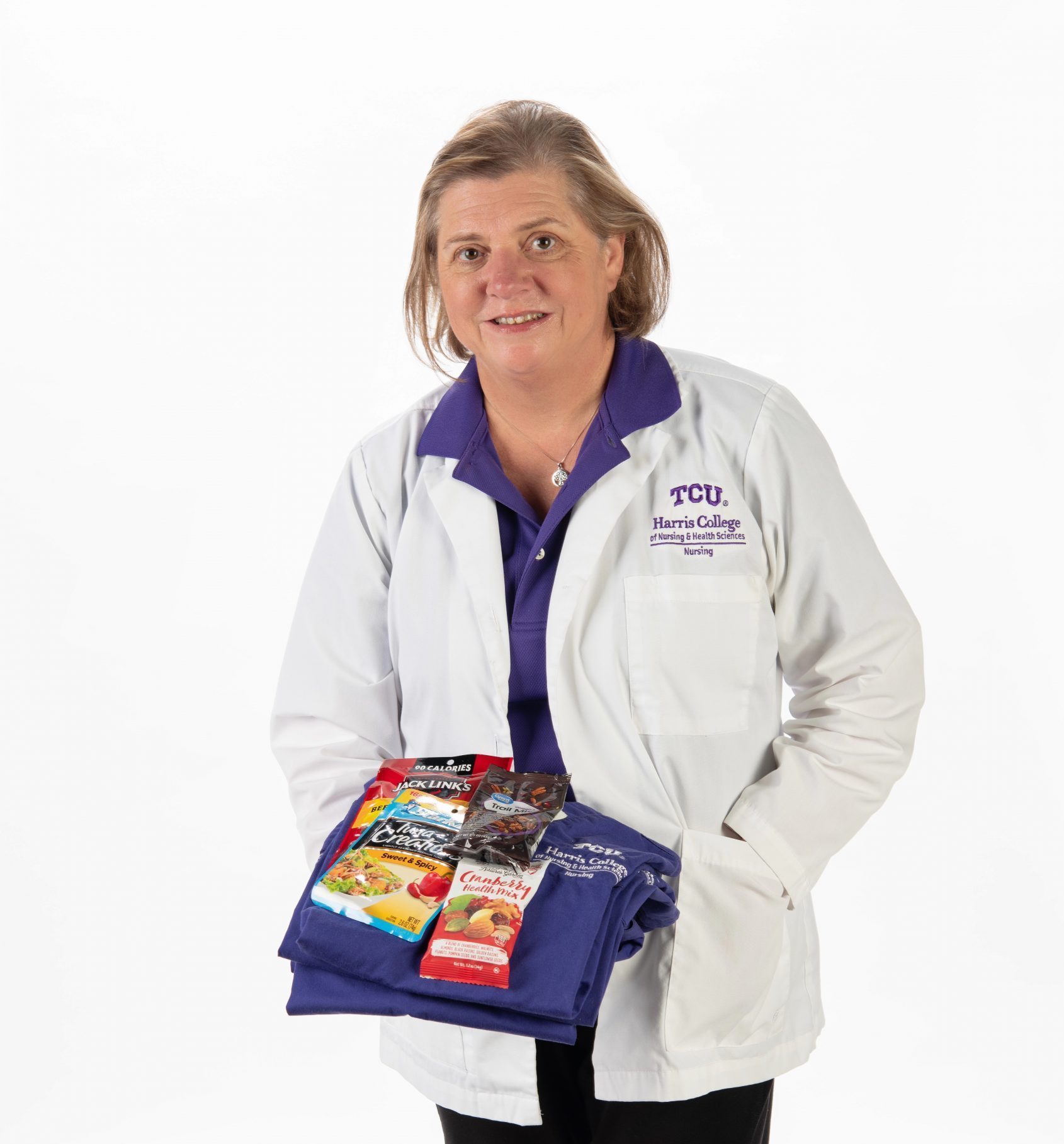 Lori Borchers, TCU nursing scholar, holds folded purple scrubs topped with packaged snacks including trail mix, beef jerky and tuna.