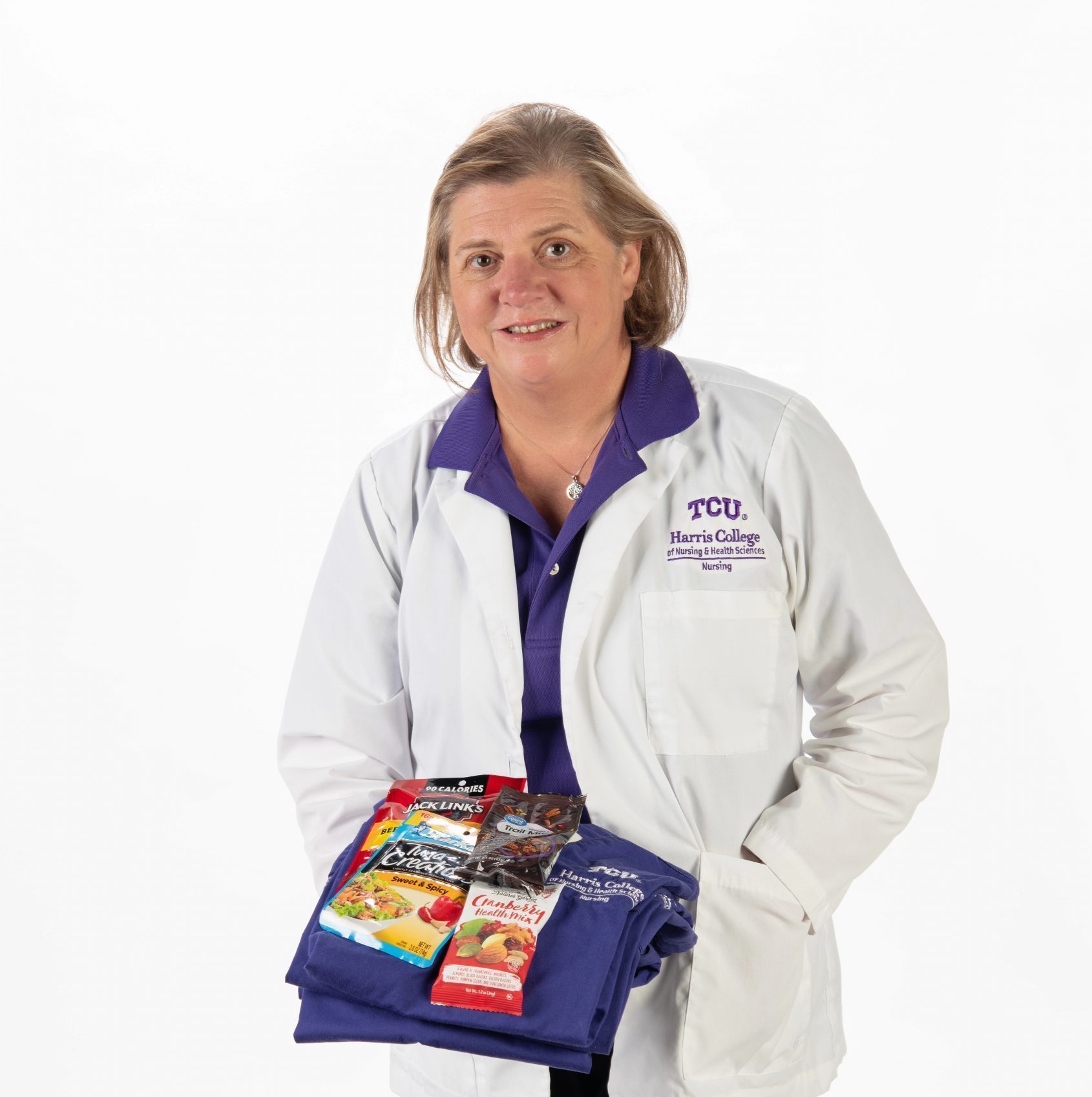 Lori Borchers, TCU nursing scholar, holds folded purple scrubs topped with packaged snacks including trail mix, beef jerky and tuna.