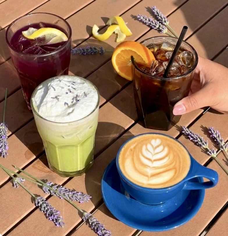 Four specialty beverages from Roots Coffeehouse arranged on a wooden table with lavender sprigs: a latte with foam art in a blue cup, a lavender matcha with whipped cream, a purple mood-changer tea with lemon, and an iced Americano with orange garnish