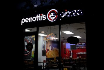 The illuminated exterior of Perrotti's Pizza at night, its white-and-red storefront sign featuring the mustachioed mascot logo, with the lit dining room and a neon pizza-slice sign visible through the front windows.