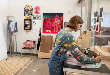 Caya Crum works at a table in her art studio, seen from behind in an embroidered denim jacket, with a large pink painting of figures in cowboy hats hanging on the wall behind her. Photo by Pilar Olivas