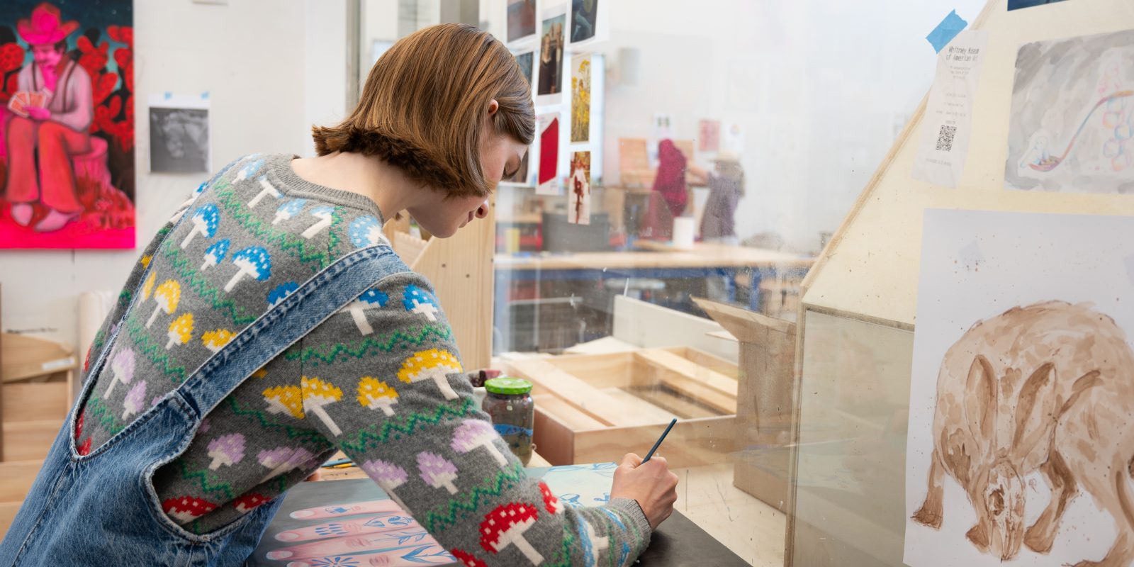 Caya Crum works at a table in her art studio, seen from behind in an embroidered denim jacket, with a large pink painting of figures in cowboy hats hanging on the wall behind her.