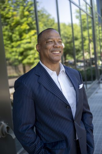 John Booty stands outside a glass-fronted building, hands in his pockets, smiling and looking off to his left. He wears a navy pinstriped suit, a white open-collar shirt and a white pocket square. Trees and a paved walkway are visible behind him.
