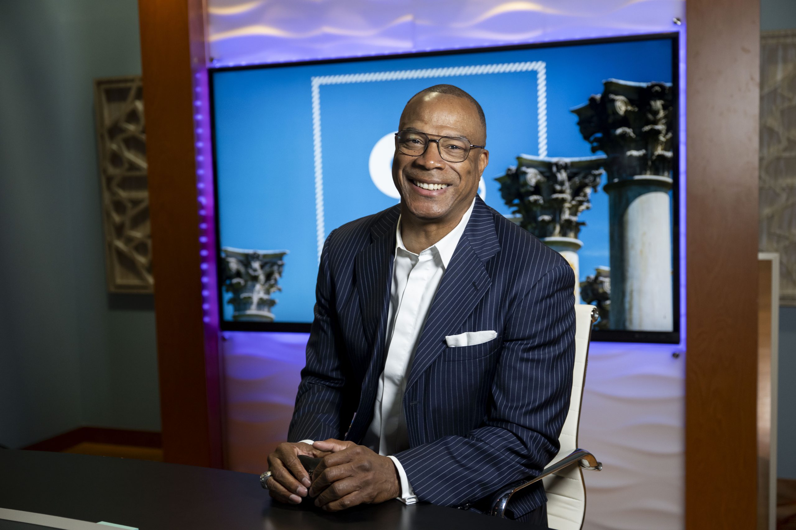 John Booty, wearing glasses, a navy pinstriped suit and a white open-collar shirt, sits at a dark desk with his hands clasped, smiling at the camera. Behind him a large screen displays a blue graphic with ornate stone columns, edged by purple lighting.