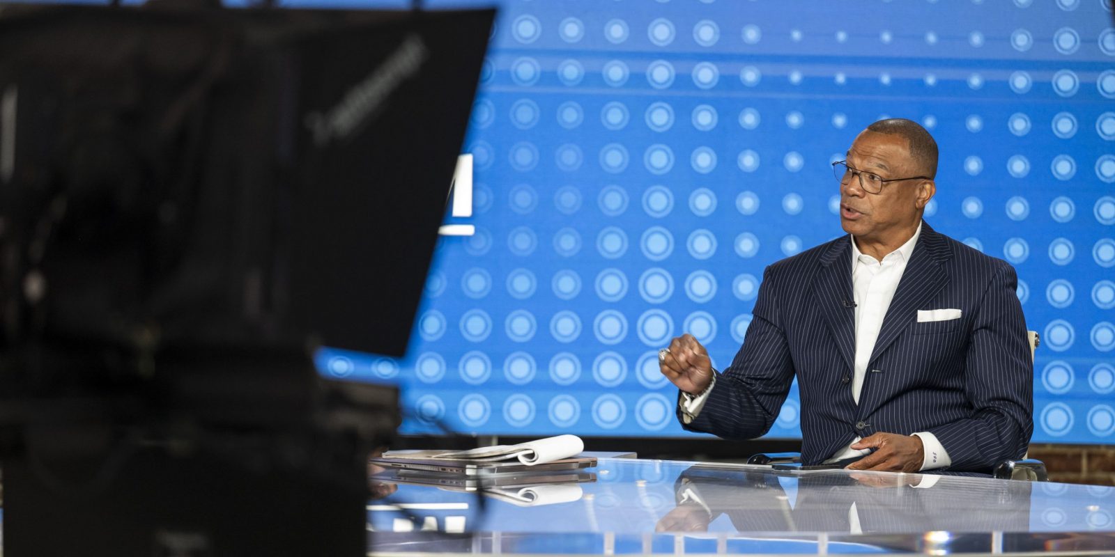 John Booty, wearing glasses and a navy pinstriped suit with a white pocket square, sits at a glass desk in a television studio, turned to his right mid-sentence with one hand raised. Papers and a laptop rest on the desk. A blue backdrop patterned with rows of circles fills the wall behind him, and studio cameras and monitors frame the shot in the foreground.