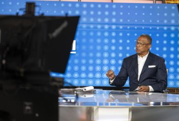 John Booty, wearing glasses and a navy pinstriped suit with a white pocket square, sits at a glass desk in a television studio, turned to his right mid-sentence with one hand raised. Papers and a laptop rest on the desk. A blue backdrop patterned with rows of circles fills the wall behind him, and studio cameras and monitors frame the shot in the foreground.