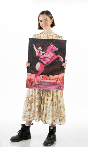 Caya Crum stands against a white background holding a painting of a cowboy on a rearing pink horse under a starry sky, with pink and red desert plants below.