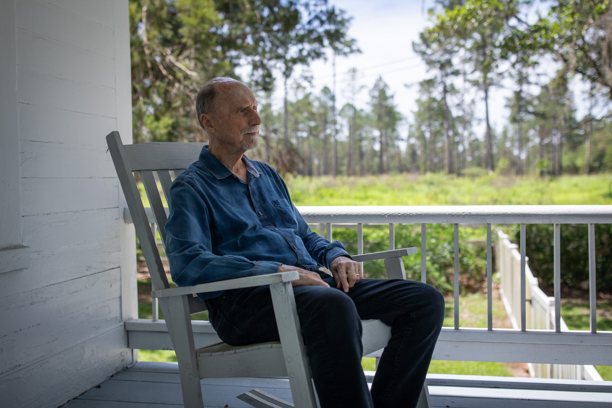 Robert Olen Butler sits in a white rocking chair on a covered porch, looking out past the railing toward pine trees and an open green field.