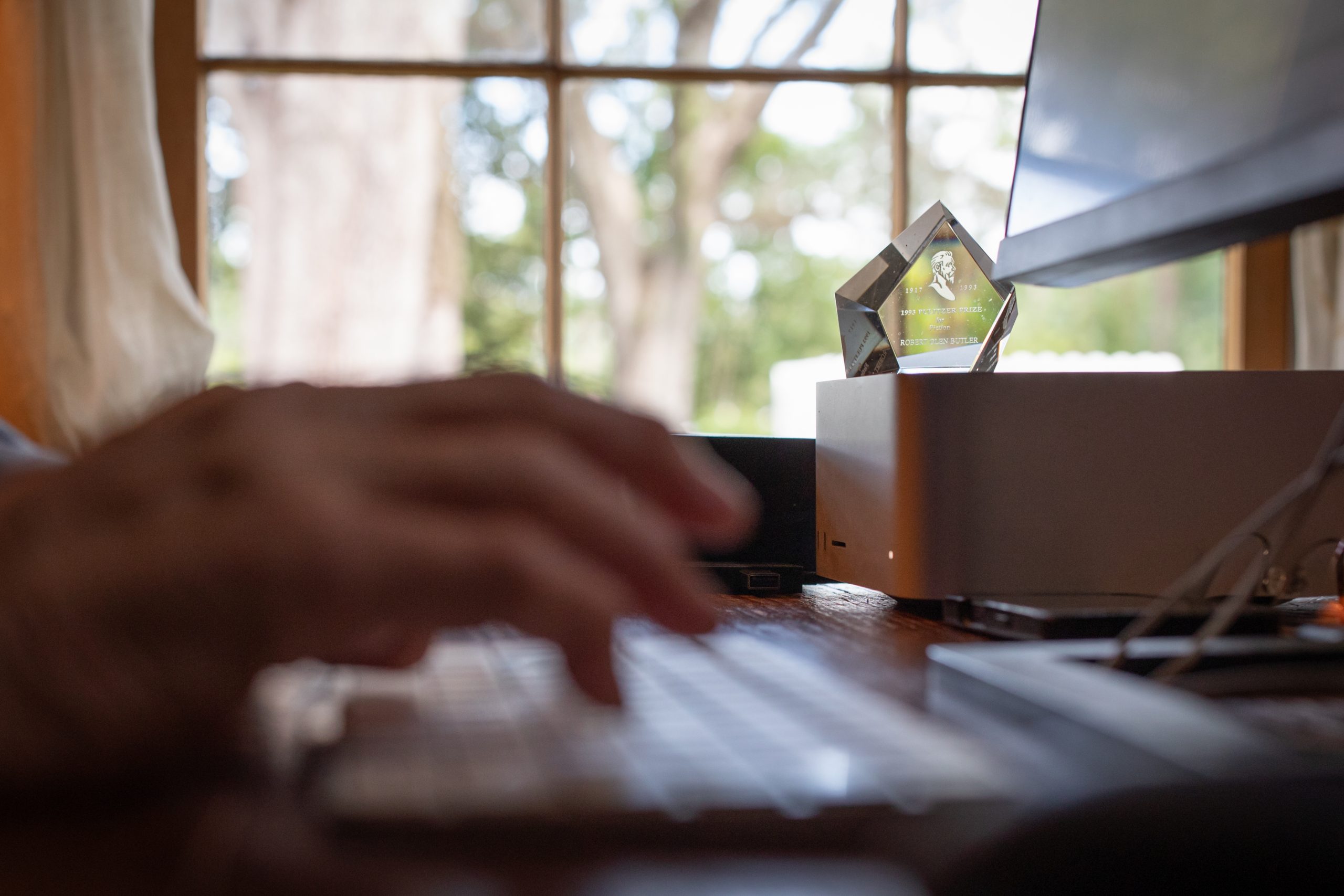 Hands rest on a laptop keyboard on a wooden desk, with a crystal award on a stand behind the screen and a bright window looking onto trees.