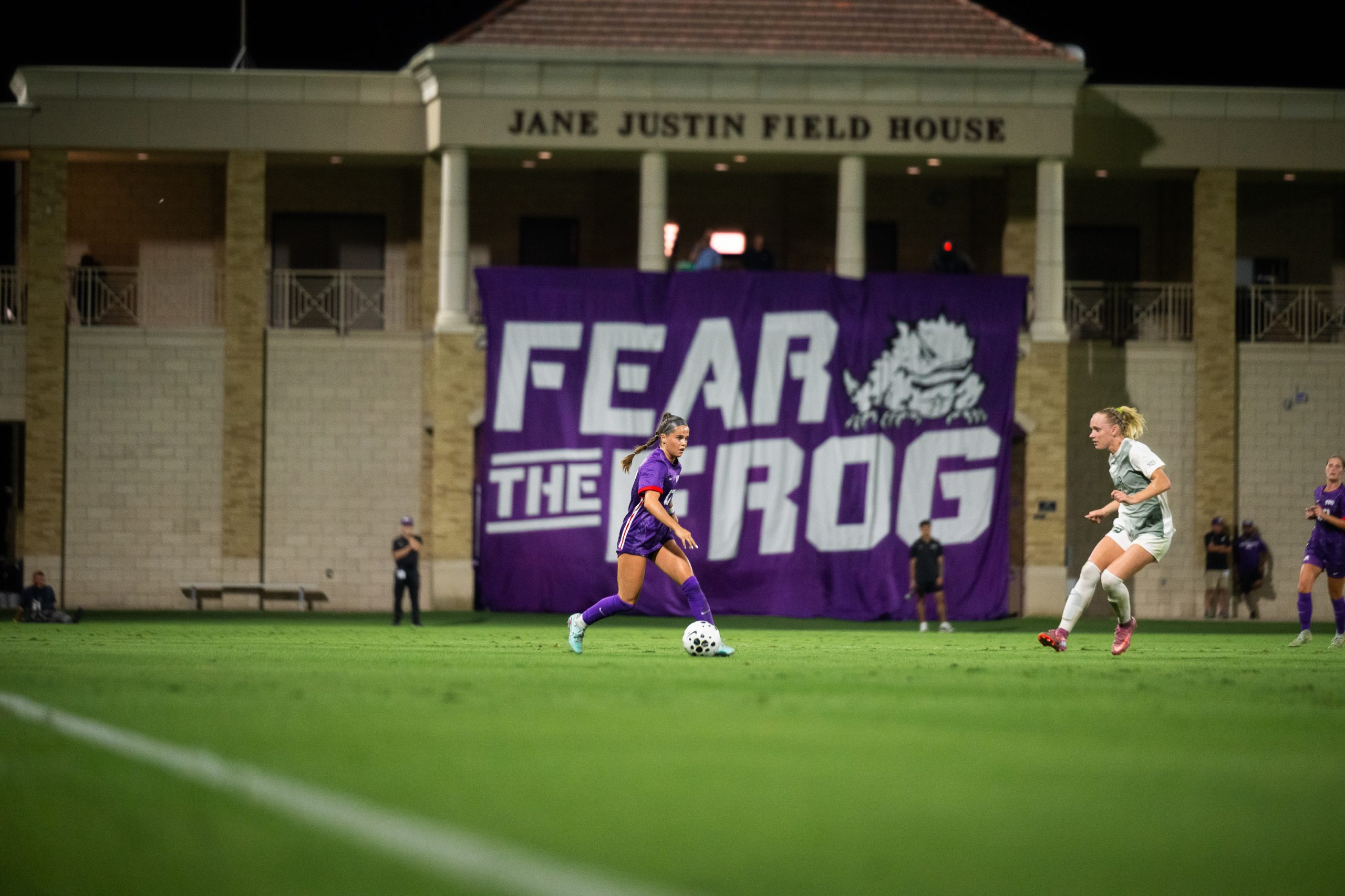 Cameron Patton moves the ball during a night match at TCU, with a Fear the Frog banner hanging from the field house behind her.