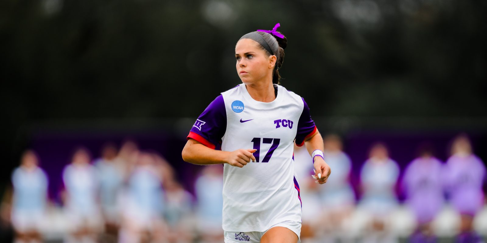 Cameron Patton on the field during a match in a white TCU uniform, with figures blurred along the sideline behind her.