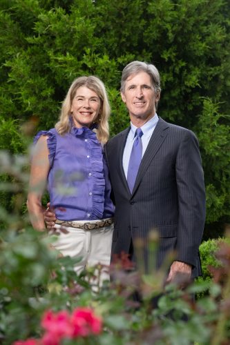 Kate Lykes and Jim Lykes stand together and smile, with dense greenery behind them and flowering plants in the foreground.