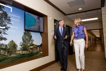 Jim and Kate Lykes walk down a hallway lined with large wall displays. Jim gestures with both hands as Kate looks toward him.