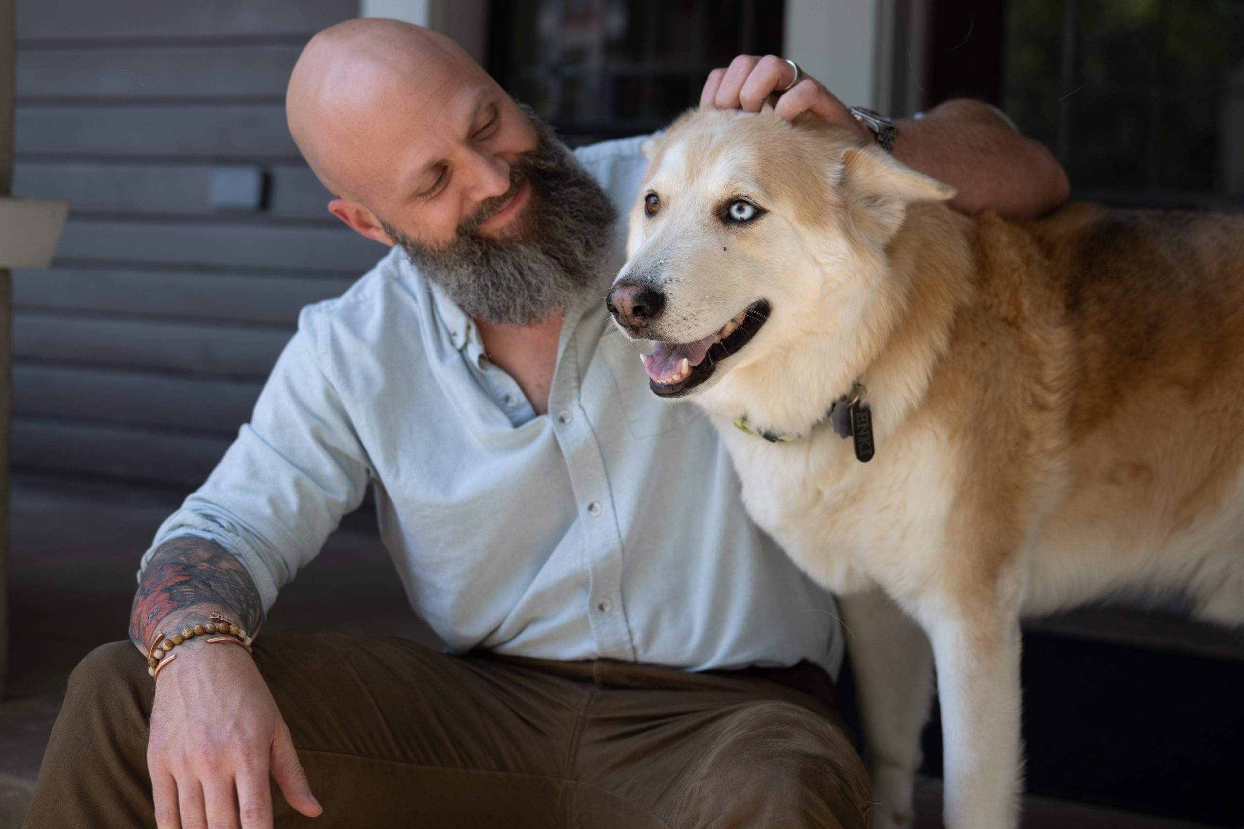 A bald, bearded man in a light blue shirt sits on a porch step, resting his hand on the head of a large cream-colored dog.