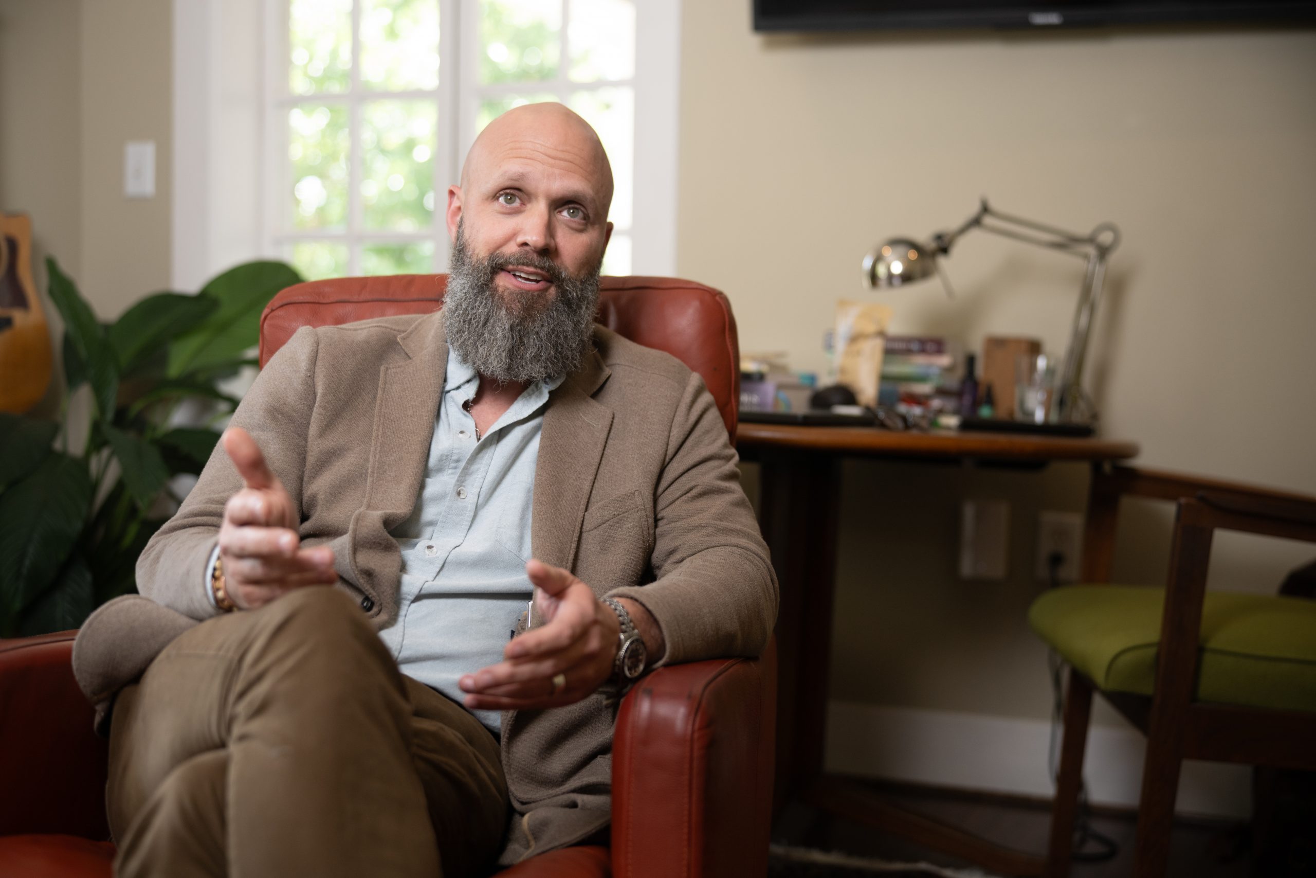 A bald, bearded man in a tan blazer sits in a red leather armchair, both hands raised mid-gesture as he speaks.