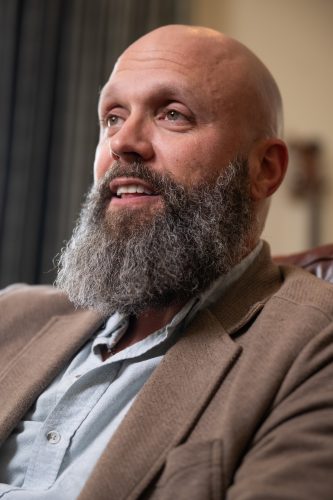 A bald man with a long gray beard sits in a leather chair, wearing a light blue button-down shirt under a tan blazer, looking off-camera and speaking.