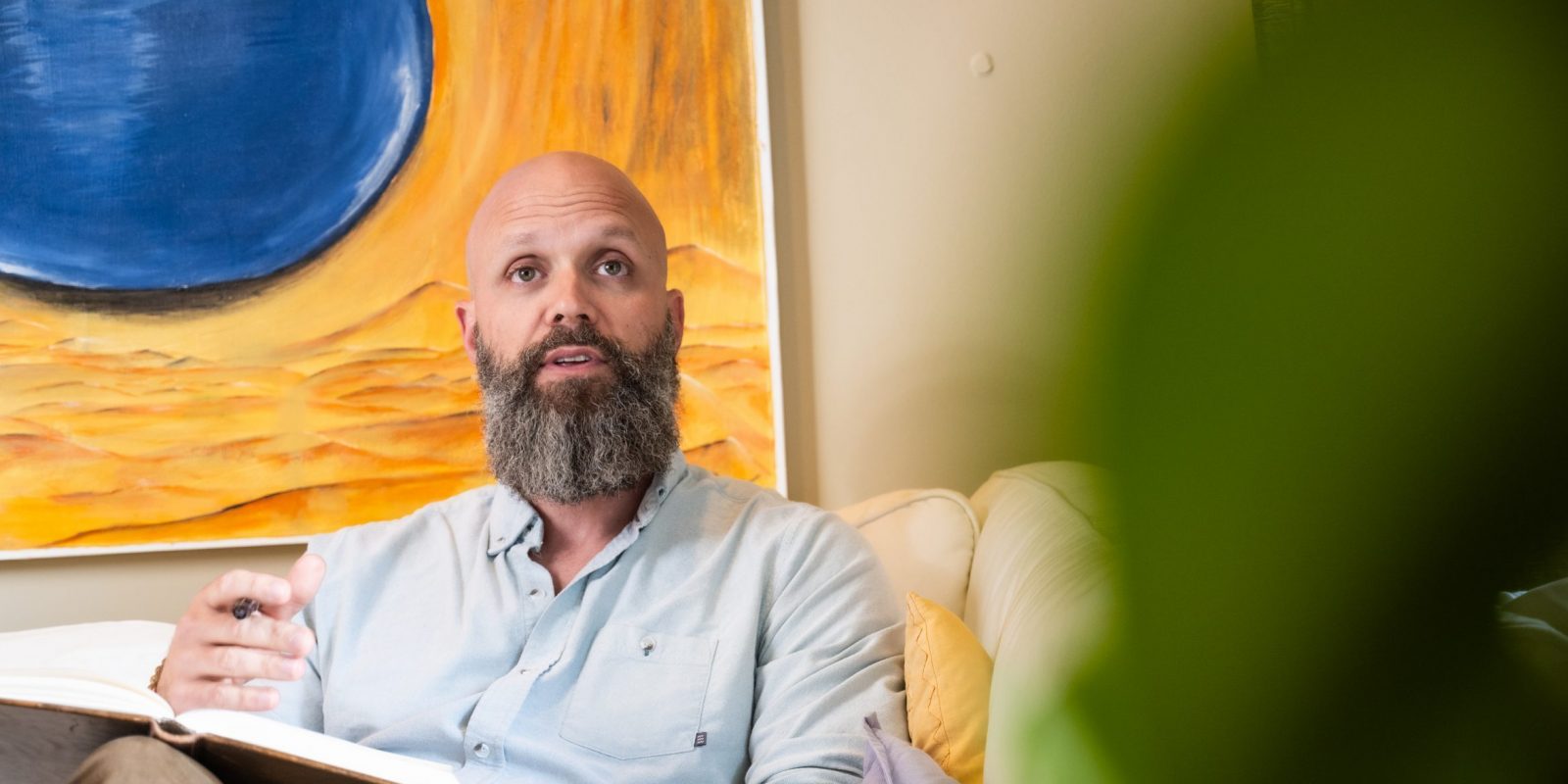 John Price sits on a couch holding an open book, one hand raised mid-gesture, with a large abstract painting on the wall behind him.