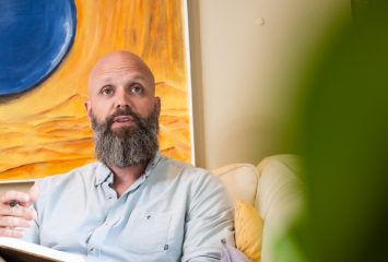 John Price sits on a couch holding an open book, one hand raised mid-gesture, with a large abstract painting on the wall behind him.