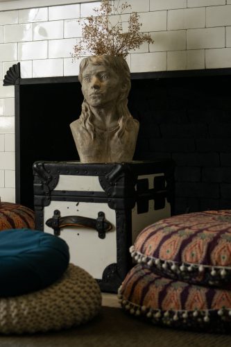 A planter shaped like a human bust, sprouting dried stems, sits on a dark wooden stand against a white tile wall, with woven floor cushions nearby.