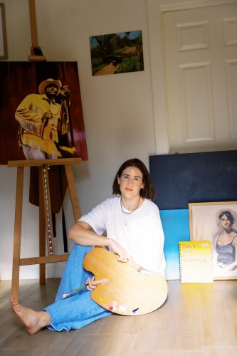Lauren Horn sits on a studio floor holding a wooden palette and brush, surrounded by canvases and an easel.