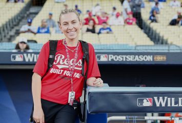 Kate Woolson stands beside a padded dugout rail marked MLB Wild Card in a red Reds T-shirt, carrying a camera and a backpack, with stadium seats and spectators behind her.