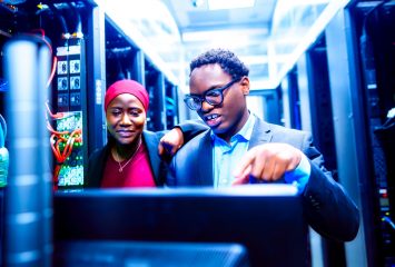 Diarra Niang and Happy Herman stand together in a server room, looking at a monitor as Herman points toward the screen. Racks of networking equipment and cabling line the aisle behind them, lit in blue.