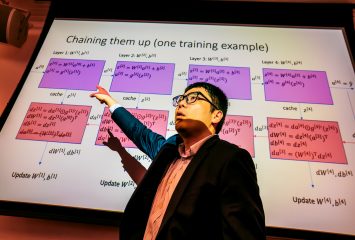 Bingyang Wei, in glasses and a dark blazer, points up at a projection screen while speaking. The slide, titled ‘Chaining them up (one training example),’ shows a neural network diagram with four layers of equations in purple and red boxes connected by arrows.