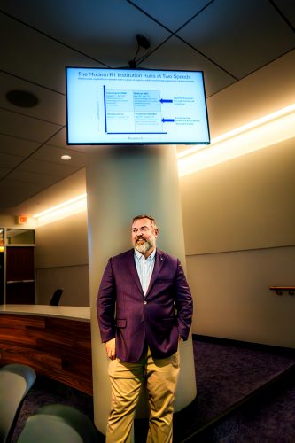 Reuben F. Burch V, in a purple blazer, checked shirt and khakis, stands against a pillar in a TCU lecture hall, looking off to the side. A wall-mounted screen above him displays a slide titled ‘The Modern R1 Institution Runs at Two Speeds,’ showing a four-quadrant chart plotting research against development.
