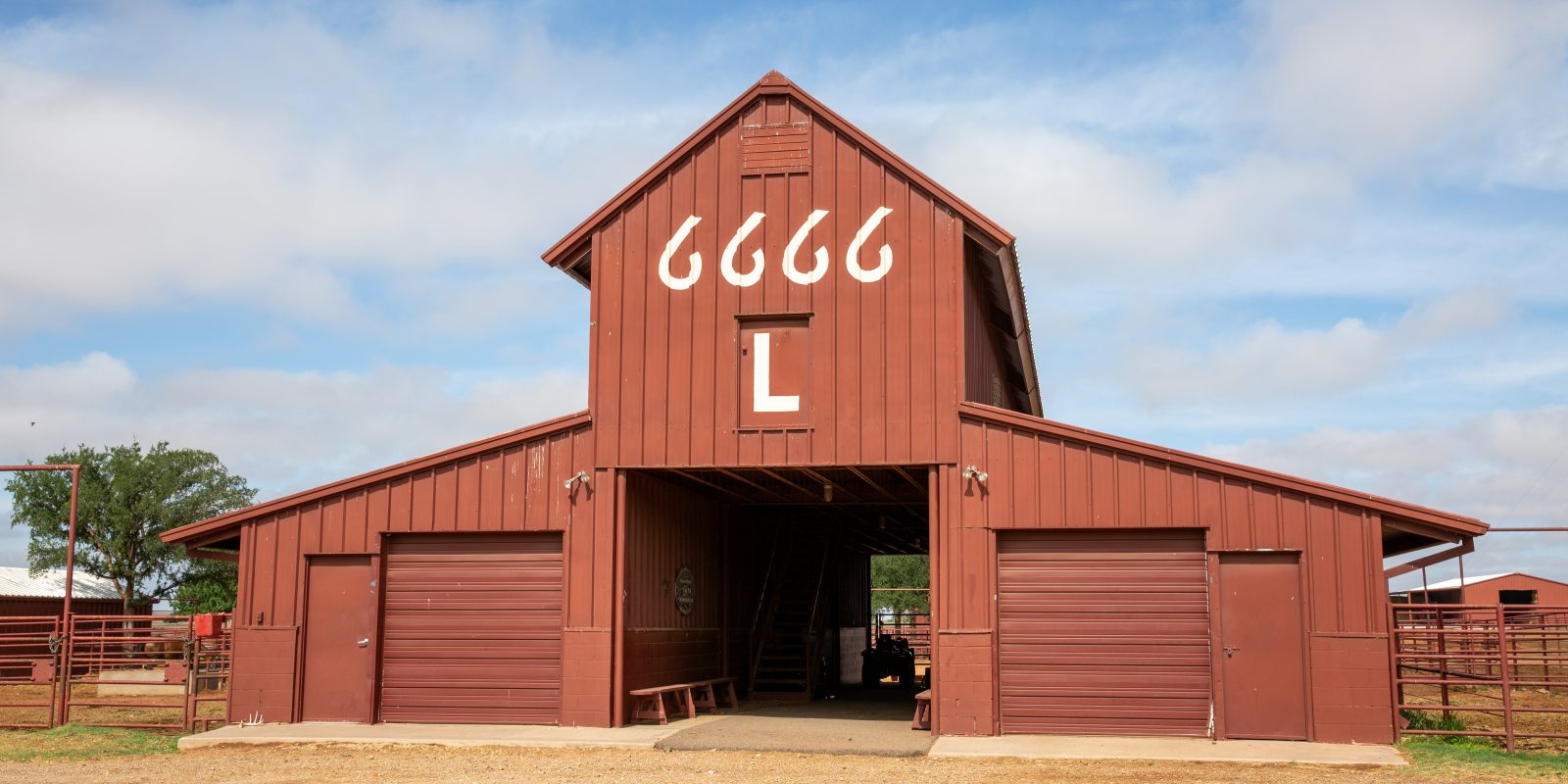 A dark red building with a peaked roof section and an open central passage, with four stylized sixes and the letter L painted above the entrance and metal fencing on either side.