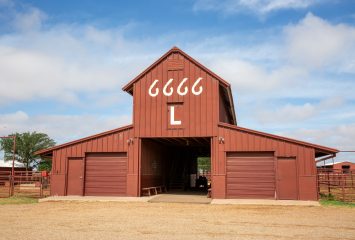 A dark red building with a peaked roof section and an open central passage, with four stylized sixes and the letter L painted above the entrance and metal fencing on either side.