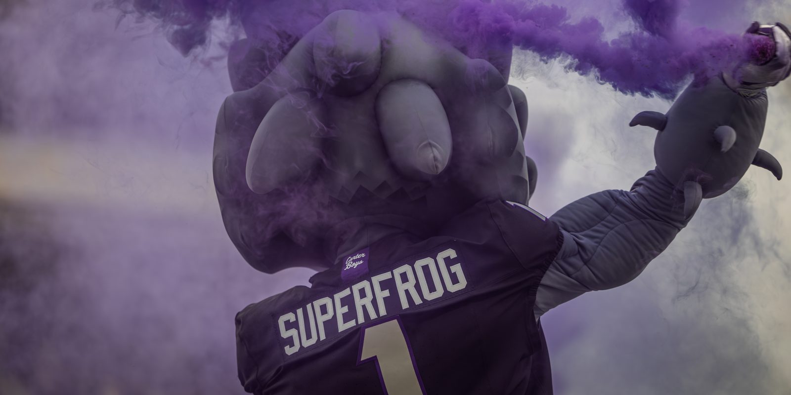 Purple smoke billows around a costumed horned frog mascot photographed from behind, wearing a jersey reading SuperFrog and the number 1, one arm raised holding a smoke device.