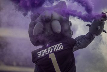 Purple smoke billows around a costumed horned frog mascot photographed from behind, wearing a jersey reading SuperFrog and the number 1, one arm raised holding a smoke device.