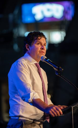 Daniel W. Pullin speaks into a microphone at a clear lectern, wearing a white dress shirt with sleeves rolled and a purple striped tie. An illuminated TCU logo glows on a screen in the dark background behind him.