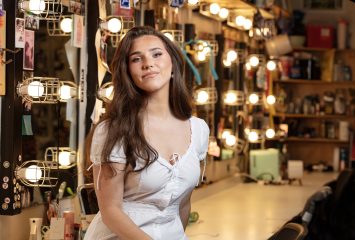 Jackie Raye sits on a dressing room counter in front of lighted mirrors.