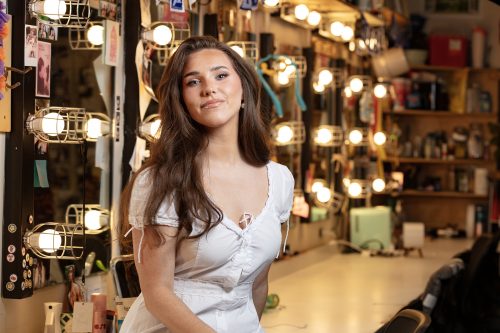 Jackie Raye sits on a dressing room counter in front of lighted mirrors.