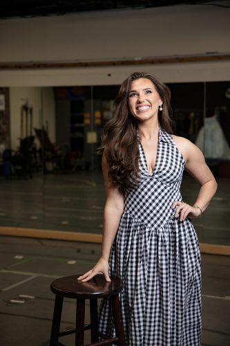 Jackie Raye smiles and looks upward, resting one hand on a stool in a mirrored rehearsal room.