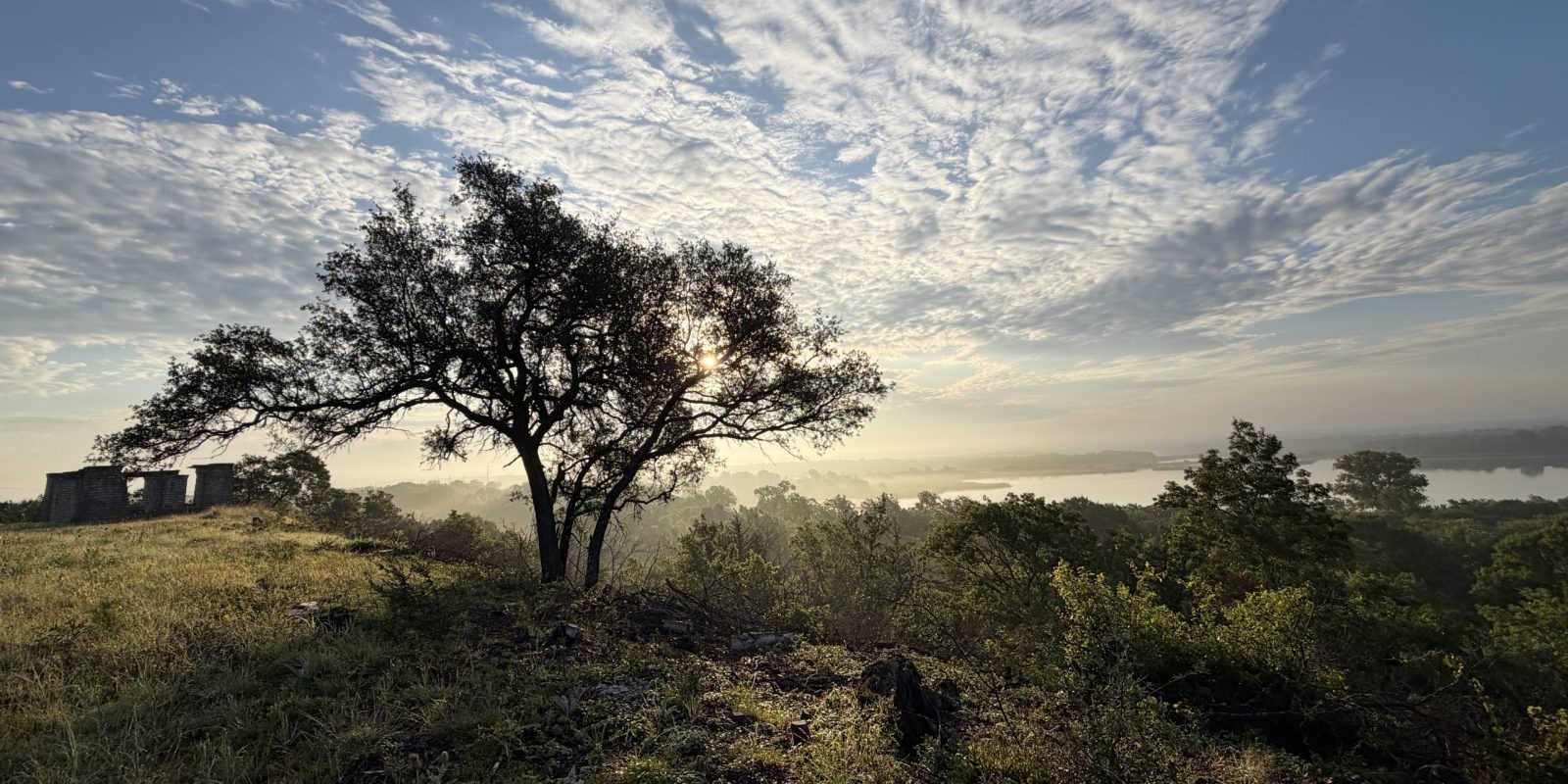 A stone structure with open archways stands on a grassy bluff beside a large tree, with mist over treetops and a lake in the distance.