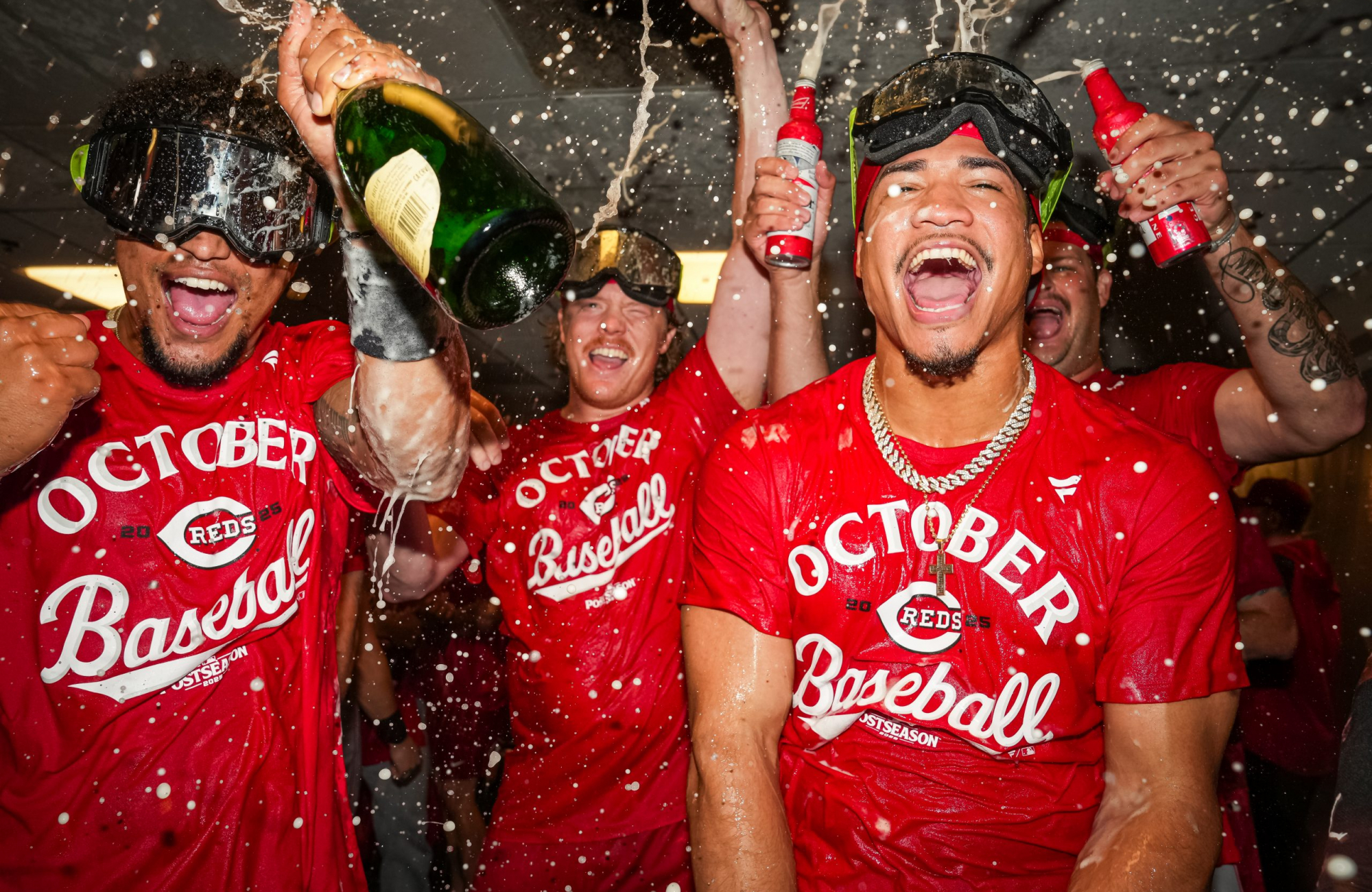 Cincinnati Reds players in red "October Baseball" postseason T-shirts celebrate in the clubhouse, wearing protective goggles and spraying champagne and beer as foam fills the air around them.