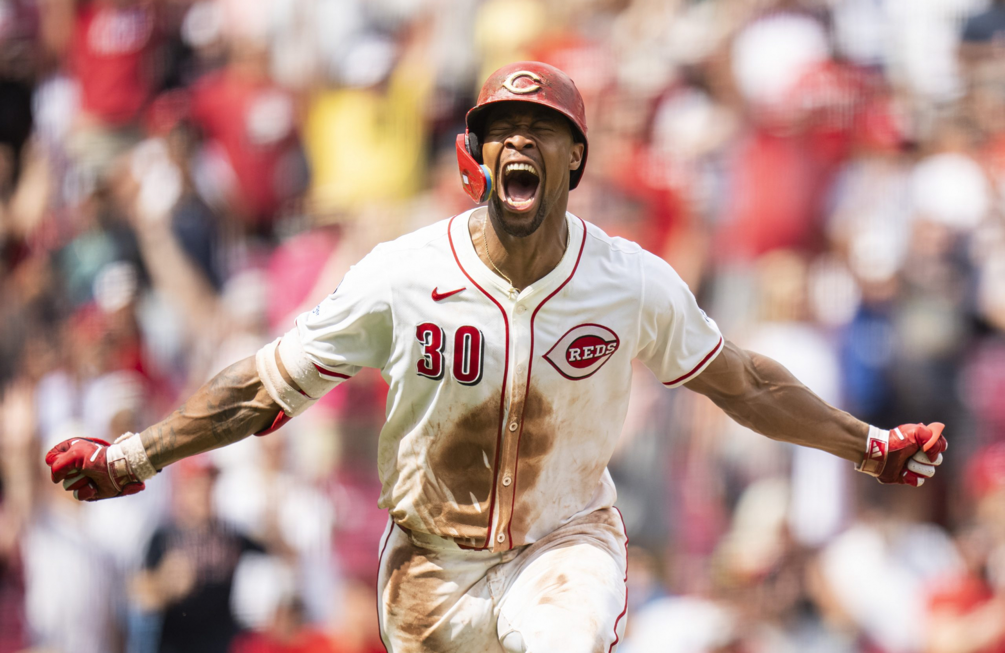 Will Benson celebrates in a dirt-streaked white Reds uniform, arms outstretched and mouth open, with a blurred crowd behind him.