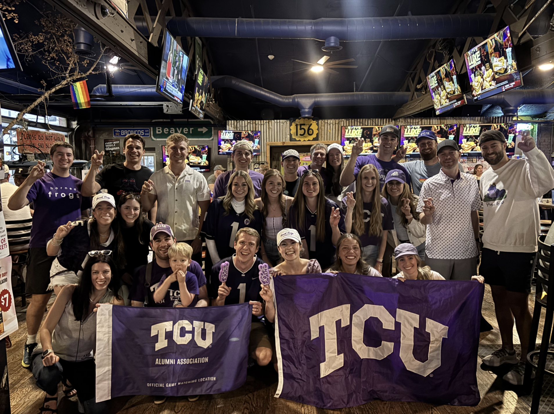 A large group of TCU fans in purple gathers for a photo inside a sports bar, holding two purple TCU flags, one reading “TCU Alumni Association, Official Game Watching Location.”