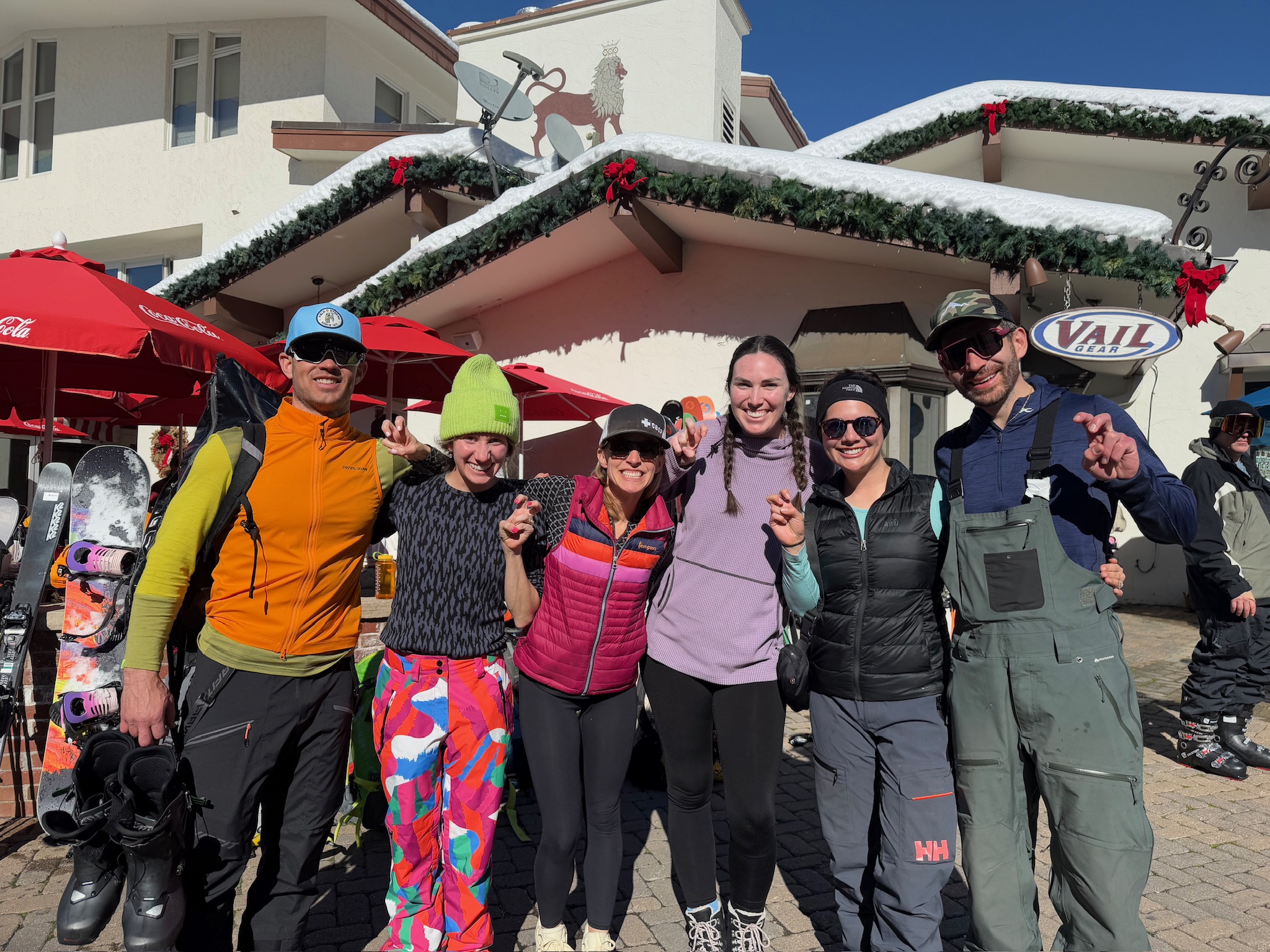 Six people in ski gear stand together outdoors on a sunny day, several making a hand gesture toward the camera, with a snow-covered chalet and skis and snowboards propped nearby.