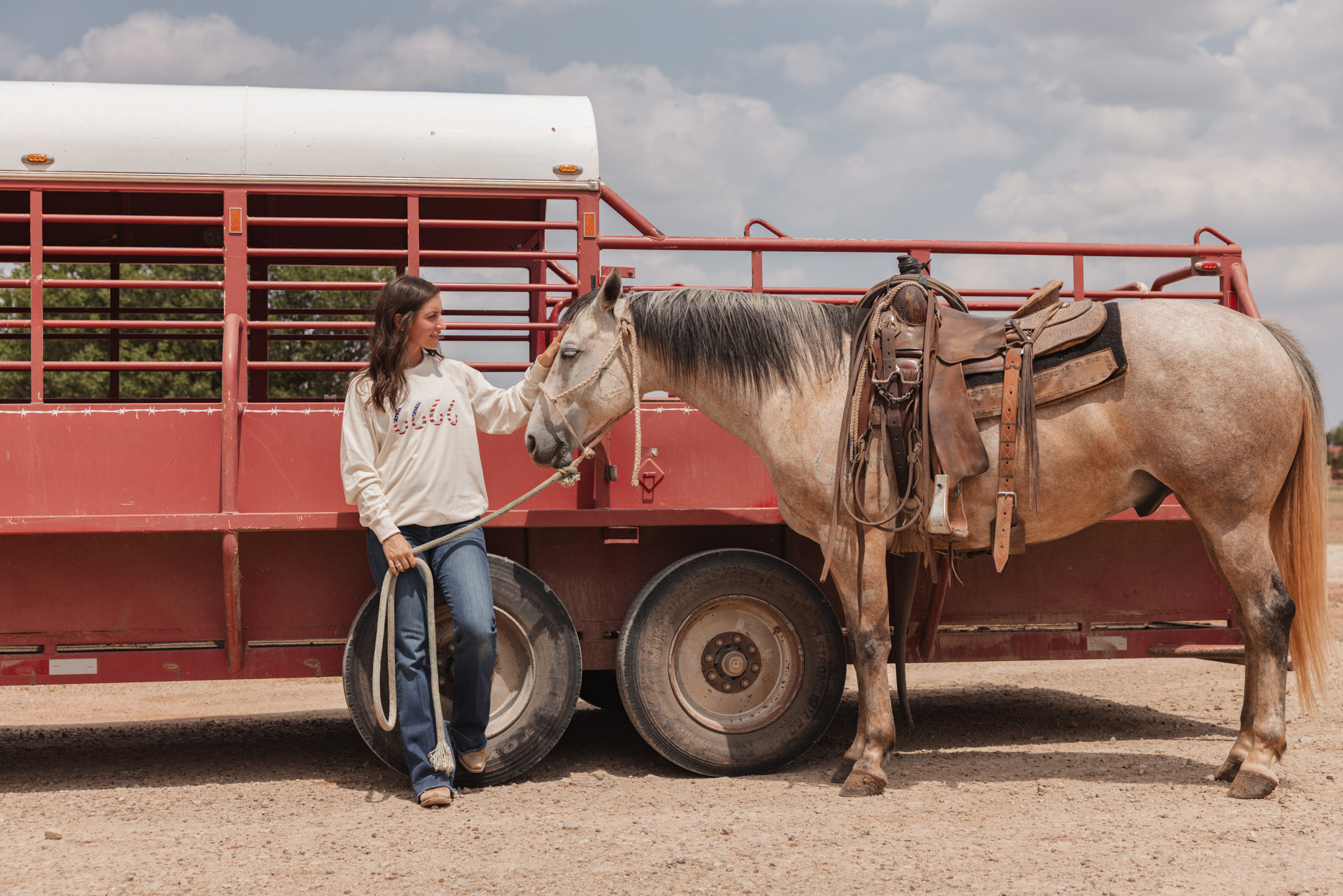 A person in a cream sweatshirt and jeans stands beside a saddled gray horse, holding a lead rope, in front of a red livestock trailer on dry ground.