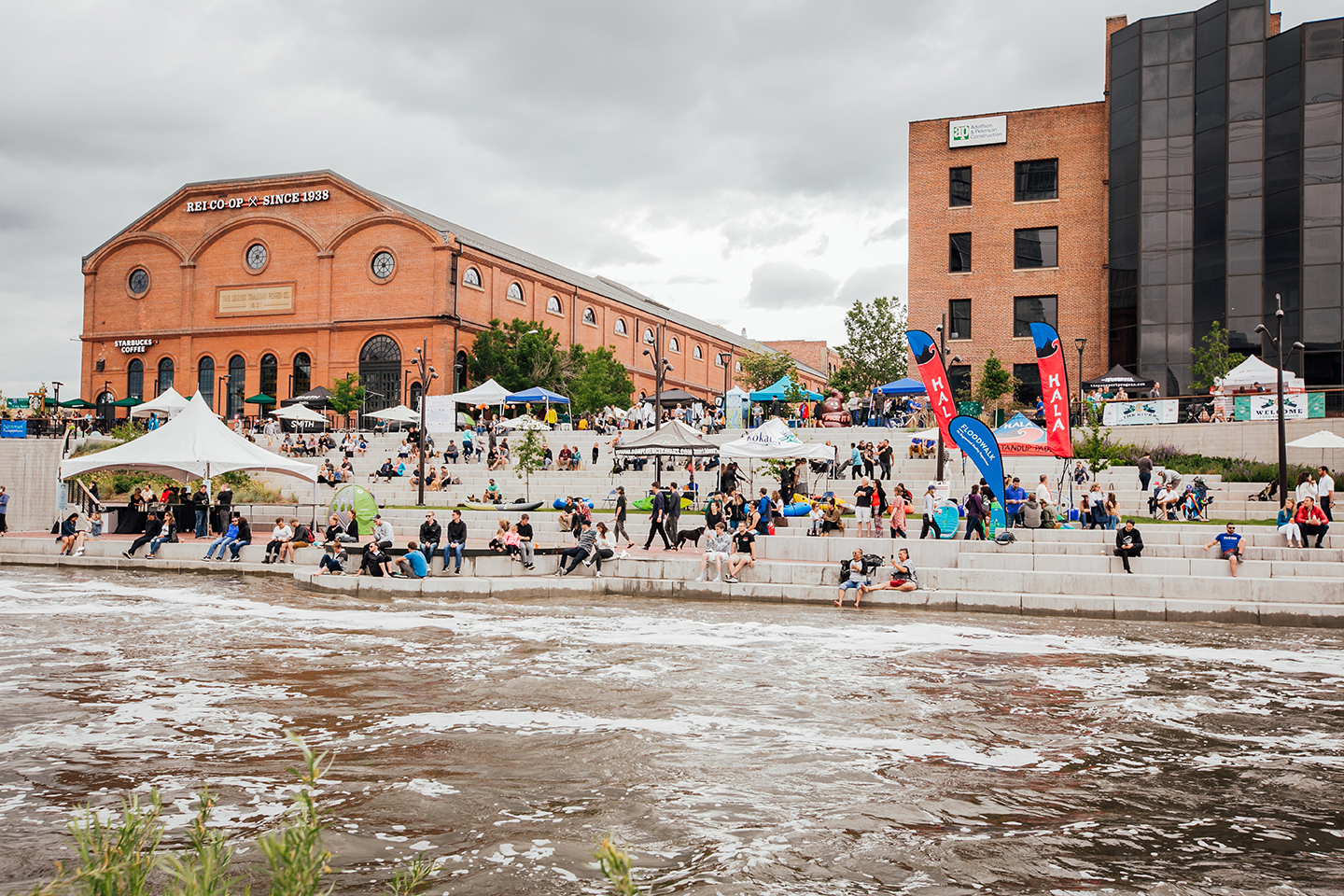 Crowds gather on tiered concrete steps along a river during an outdoor festival, with vendor tents and a historic brick REI Co-op building behind them under an overcast sky.