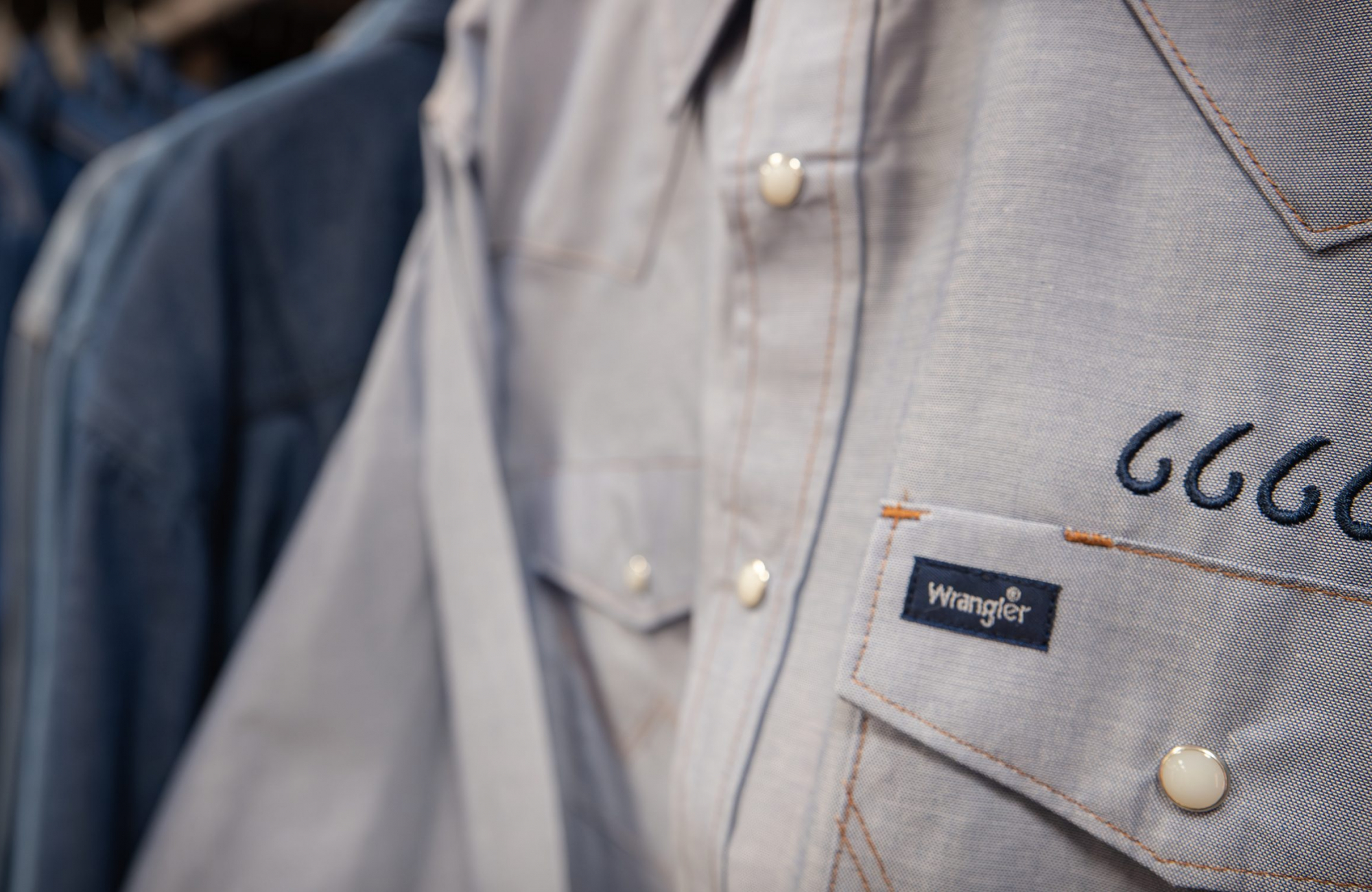 A close-up of a light gray Western-style shirt on a hanger, with pearl snap buttons, orange contrast stitching, a Wrangler label on the chest pocket and four cursive sixes embroidered in navy beside it. Blue denim garments hang blurred behind it.