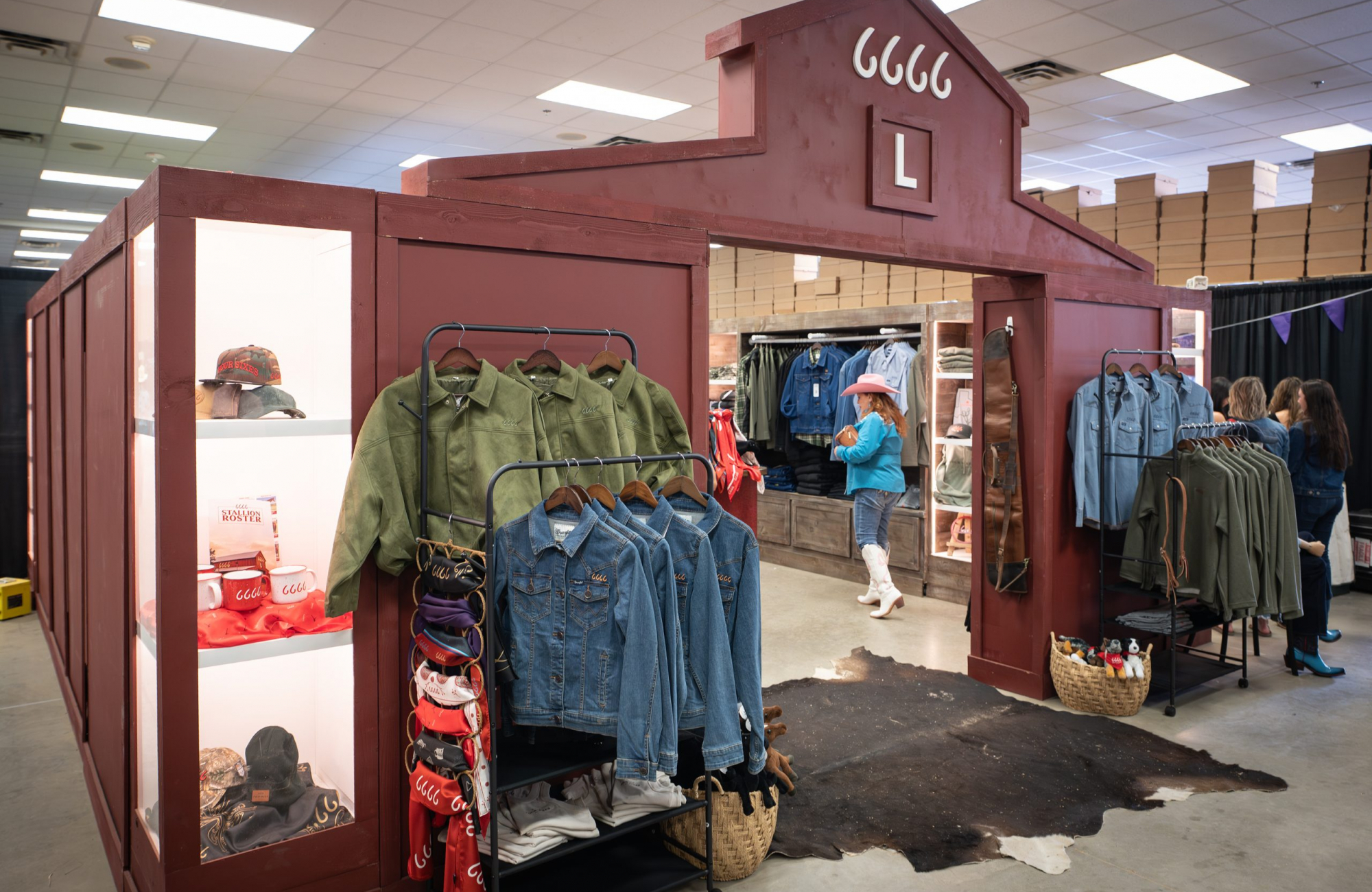 A freestanding retail booth built to look like a barn, painted dark red, with four cursive sixes and a large letter L on the gabled front. Racks of green suede jackets and blue denim jackets stand outside the entrance beside a display case holding caps, enamel mugs and a red scarf. A cowhide rug lies on the floor at the entrance, and more clothing is visible inside.