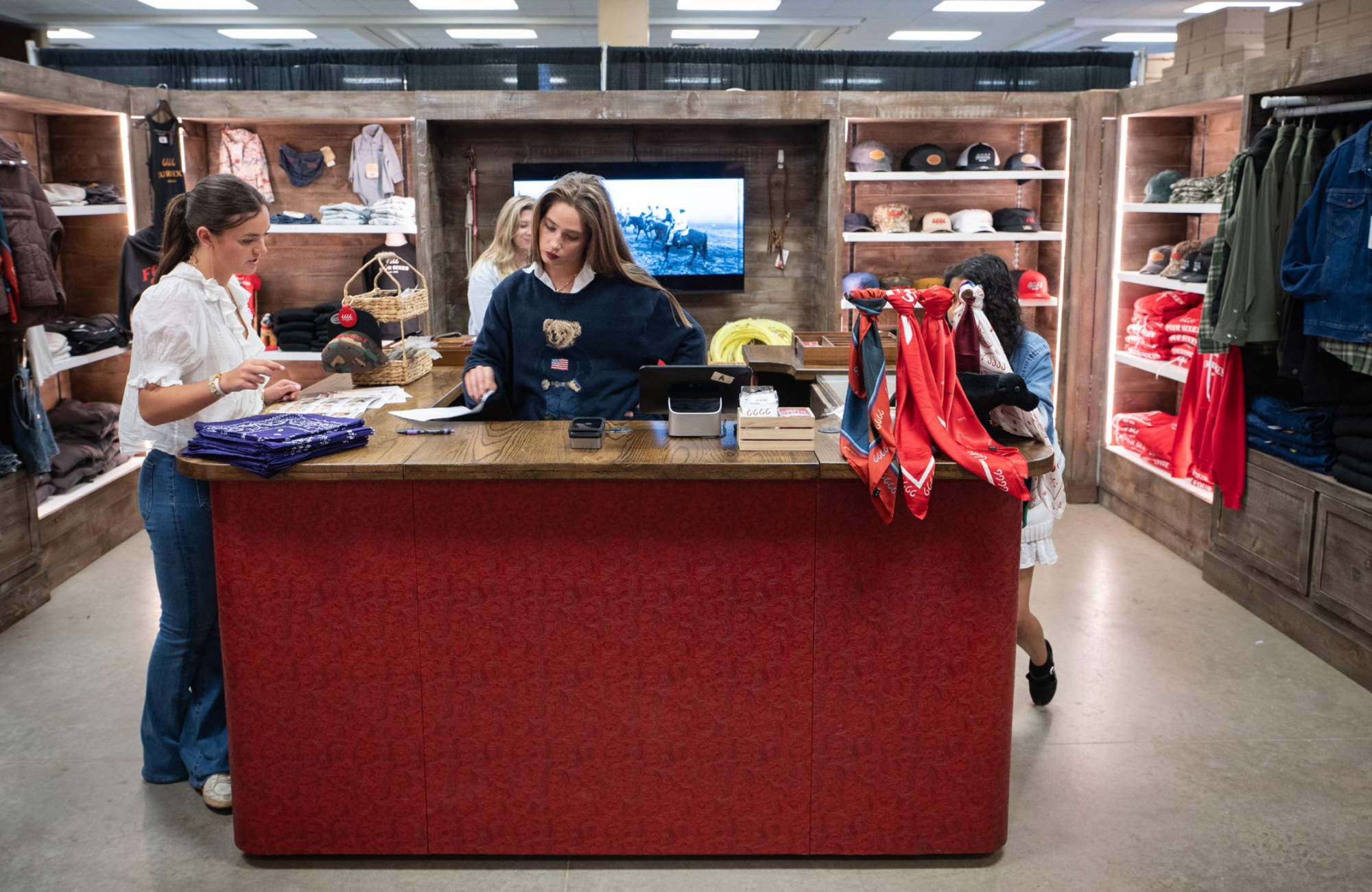 Two people work behind a large red-paneled counter in a retail space lined with wooden shelving. Folded bandanas, boxes and a tablet sit on the counter; red patterned scarves hang over one end. Racks and shelves of jackets, shirts, caps and folded denim fill the walls on both sides, and a screen behind the counter shows riders on horseback.