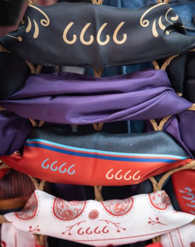 Silk scarves in black, purple, red and white are draped over a woven rope display rack, each printed with the Four Sixes brand.