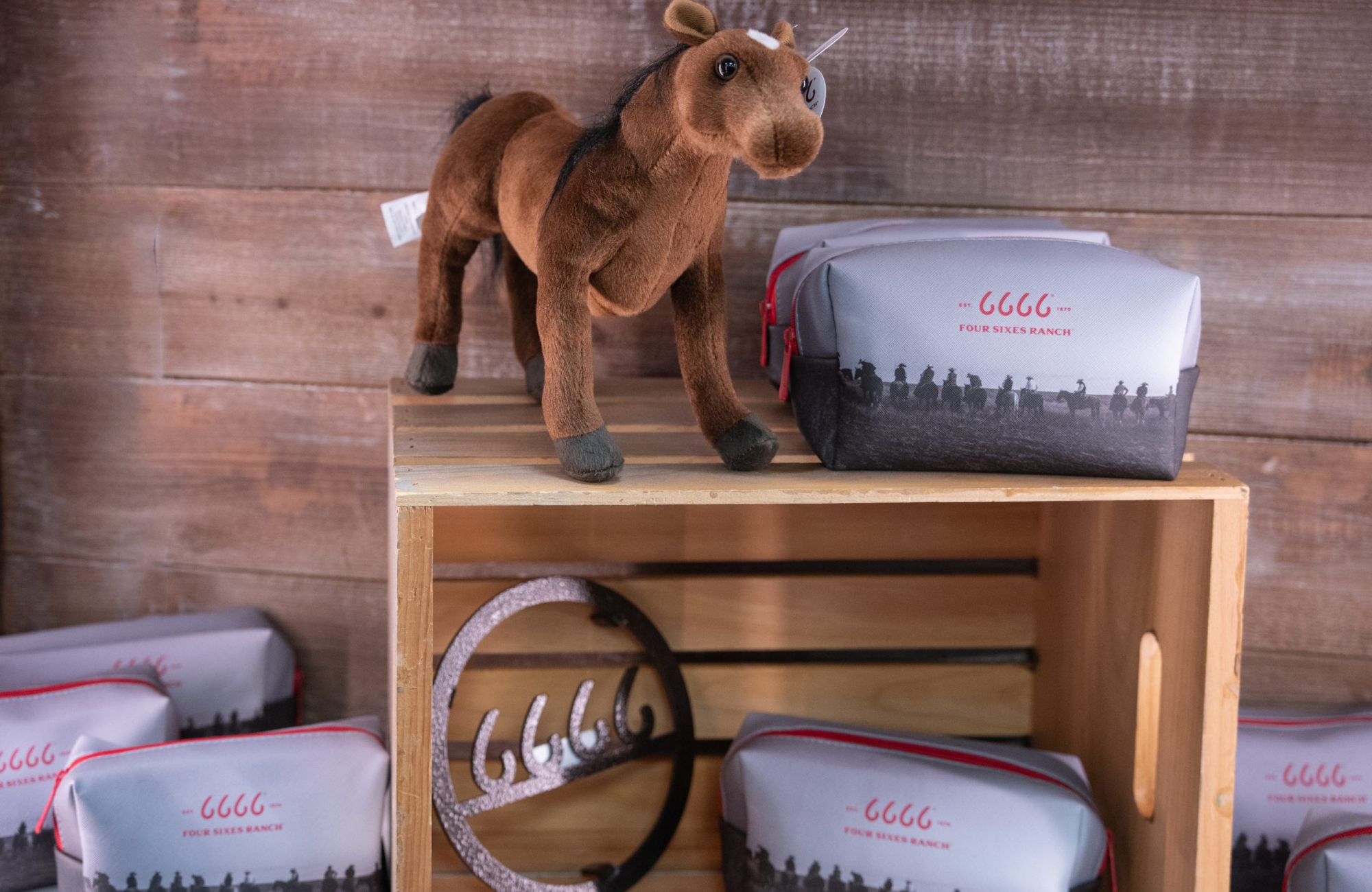 A plush toy horse stands on a wooden crate beside a gray zippered pouch printed with a red 6666 mark, the words FOUR SIXES RANCH, and a black-and-white photo of riders on horseback along the bottom. More of the same pouches are arranged below, around a metal ornament cut in the shape of four cursive sixes inside a circle.